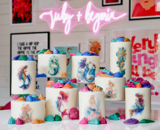 Colorful mermaid-themed cakes with decorative elements on a table, featuring the brand 'July + Begonia'.